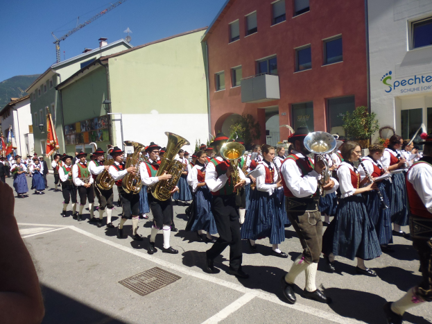 Gruppe von Menschen in traditioneller bayrischer Tracht, die eine Straße entlanggehen und Musikinstrumente spielen, einige halten Fahnen, mit Gebäuden, Pflanzen und einem Schild an der Straße, einem Hügel und einem klaren blauen Himmel im Hintergrund.