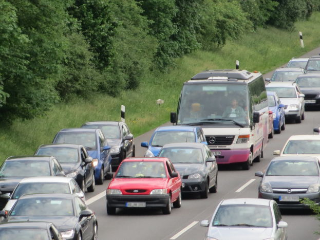 Ein Stau auf einer Autobahn mit zahlreichen Autos und einem Lieferwagen, Menschen in den Fahrzeugen, Bäume und Gras im Hintergrund.