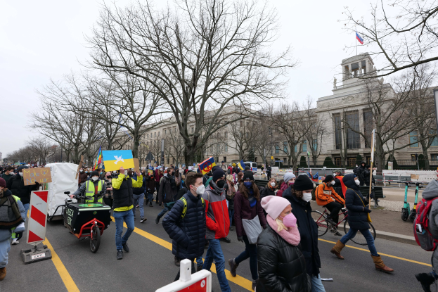 Eine große Gruppe von Menschen marschiert bei einer Demonstration auf einer Stadtstraße, einige halten Schilder und andere fahren Fahrräder, mit Bäumen und einem Gebäude im Hintergrund unter einem klaren blauen Himmel.