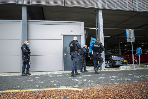 Eine Gruppe von Polizeibeamten vor einem Gebäude mit einer Tür, umgeben von Fahrzeugen, Pfosten, Brettern und verstreuten Steinen auf einem Parkplatz.