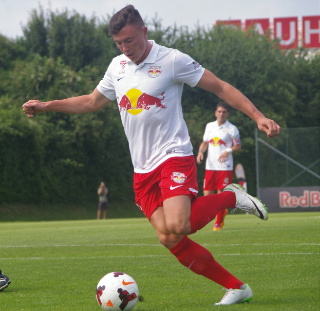 Professioneller Fußballspieler, der einen Ball auf einem Rasenfeld mit dem Hintergrund "RB Leipzig"-Schriftzug unter einem klaren blauen Himmel kickt.