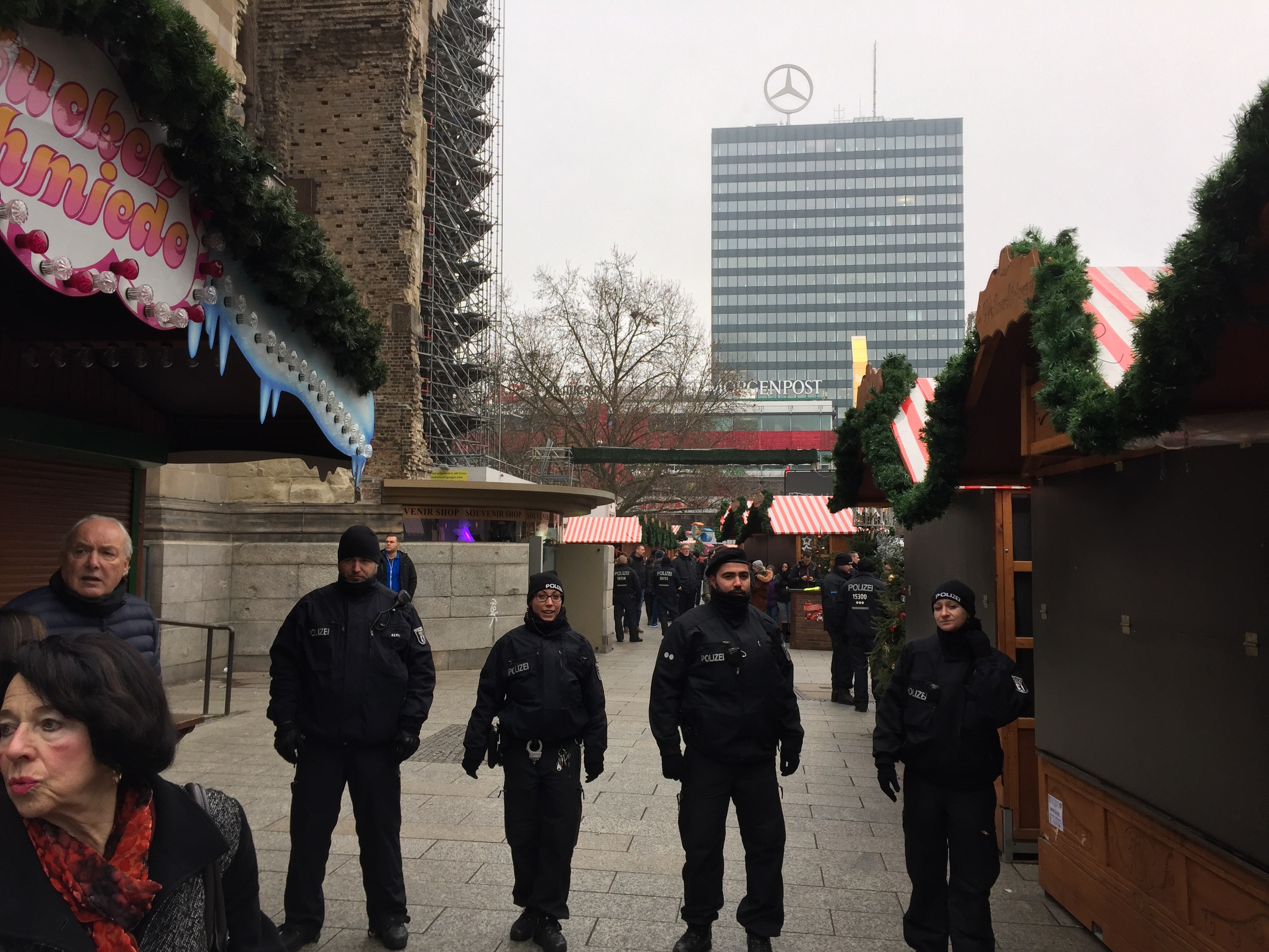 Polizeibeamte in schwarzen Uniformen und Mützen vor einem geschmückten Weihnachtsmarkt in Berlin, Deutschland, mit Gebäuden, Bäumen und einem klaren blauen Himmel im Hintergrund.