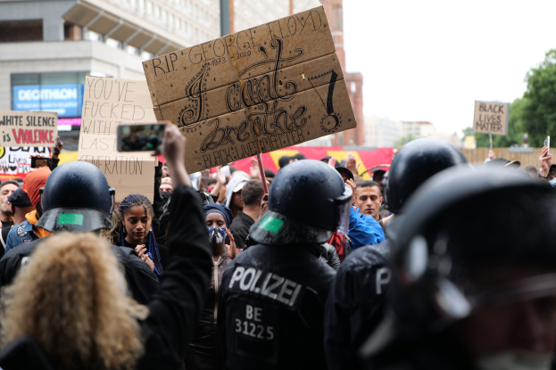 Gruppe von Menschen mit Helmen und Schildern bei einer Demonstration vor einem Gebäude, Bäumen und einem Pfahl im Hintergrund.