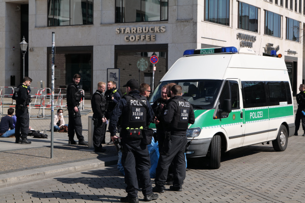 Polizisten vor einem Starbucks-Café mit einem Van auf der rechten Seite und Menschen auf der linken Seite vor einem Gebäude mit Fenstern, einem Schild, einem Laternenmast und einem Zaun.