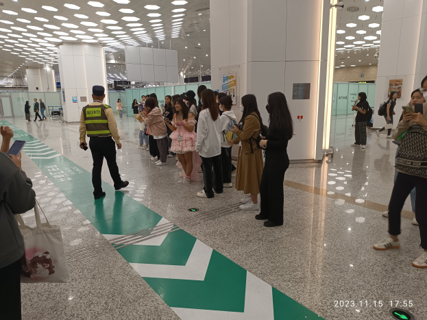 Menschen in einer Reihe an einem Flughafen-Check-in-Bereich mit Gepäck und Mobiltelefonen, mit Säulen und Deckenleuchten im Hintergrund.