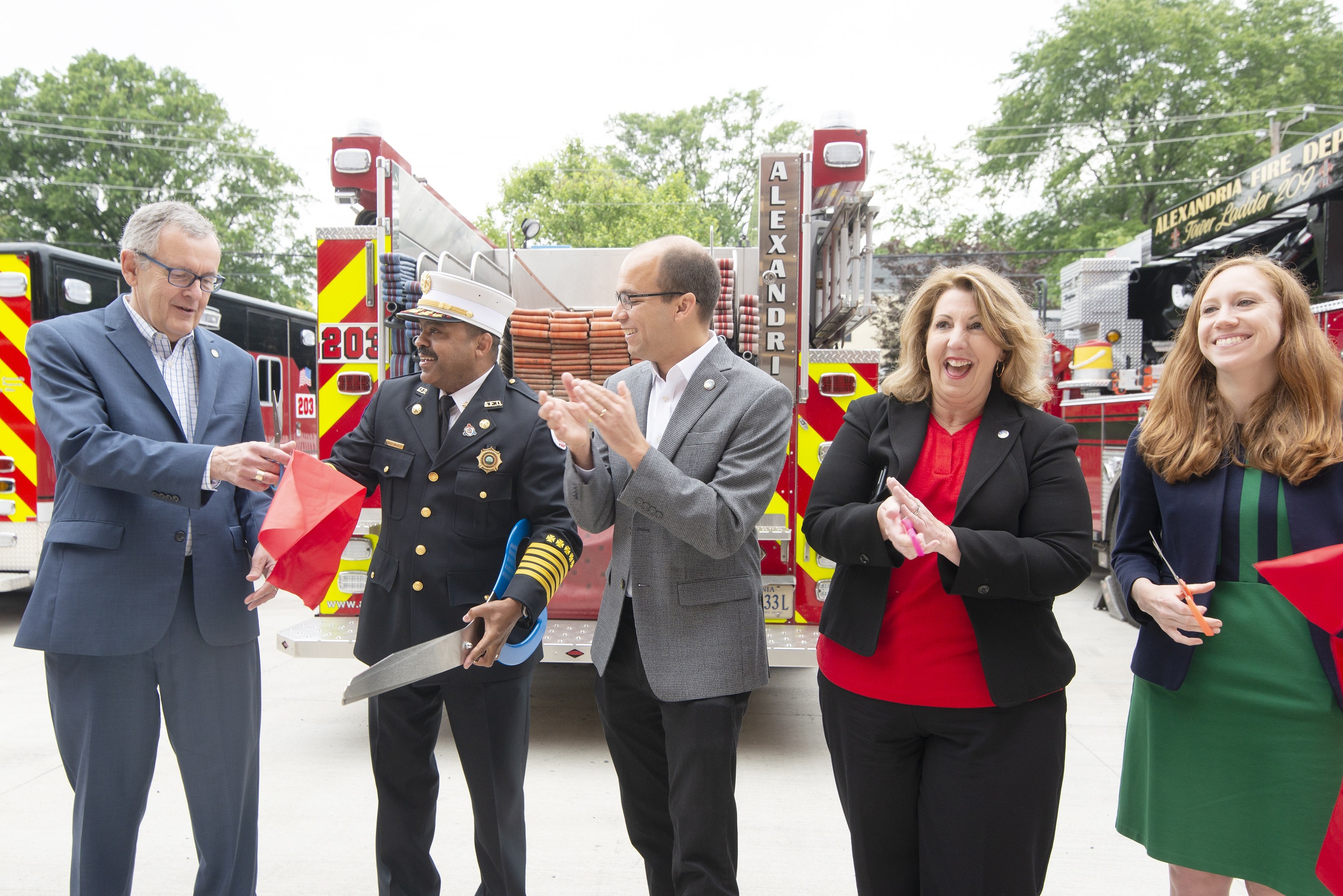 Gruppe von Menschen, die bei einer Einweihungszeremonie der Alexandria Fire Department klatschen und lächeln, mit zwei Personen, die eine Schere und ein rotes Band halten, vor einem Feuerwehrauto.