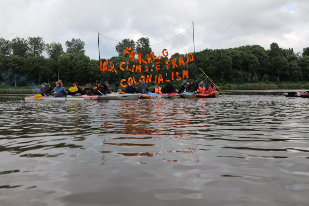 Gruppe von Menschen in Kajaks auf einem Gewässer, einige halten Paddel, mit Bäumen und einem klaren blauen Himmel im Hintergrund und einer Fahne mit Text in der Mitte.