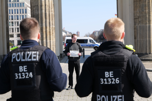 Drei Polizisten stehen vor einem Gebäude, ein Mann in der Mitte hält ein Papier, flankiert von Säulen, während Fahrzeuge, Menschen, Bäume und Gebäude den Hintergrund und den Himmel ausfüllen.