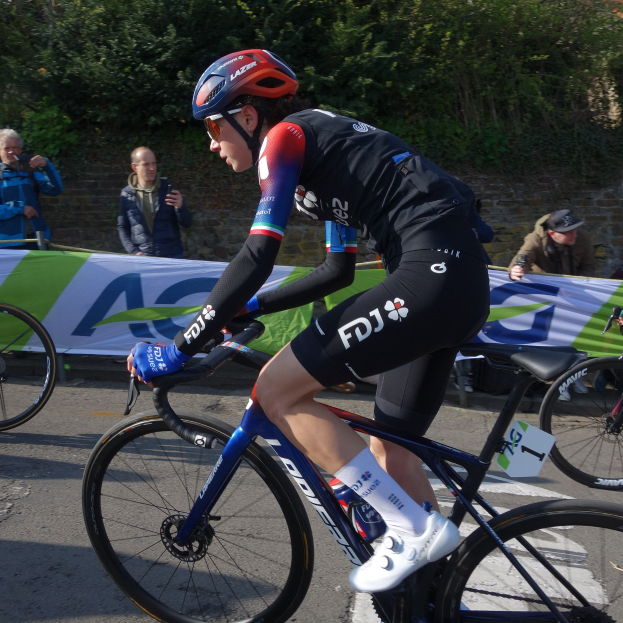 Eine professionelle Radrennfahrerin, Katie Archibald, fährt in Helm, Brille, Handschuhen und Schuhen ein Fahrrad auf einer Straße neben einer Menschenmenge. Im Hintergrund gibt es eine Wand, Bäume und ein Gebäude.