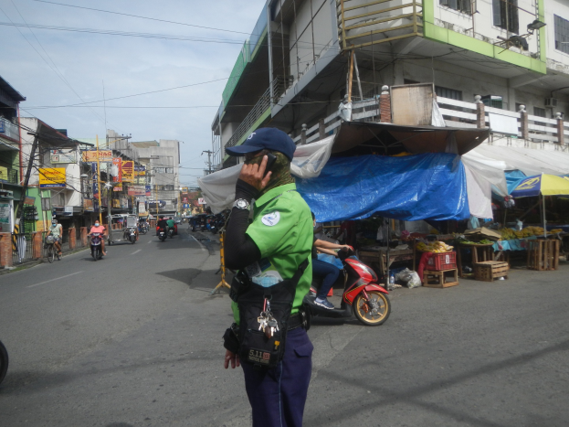 Polizist in grüner und blauer Uniform mit blauer Mütze auf einer Straße stehend, der auf einem Handy spricht, mit Fahrzeugen auf der Straße, Marktständen rechts und einem Gebäude im bewölkten Himmel im Hintergrund.