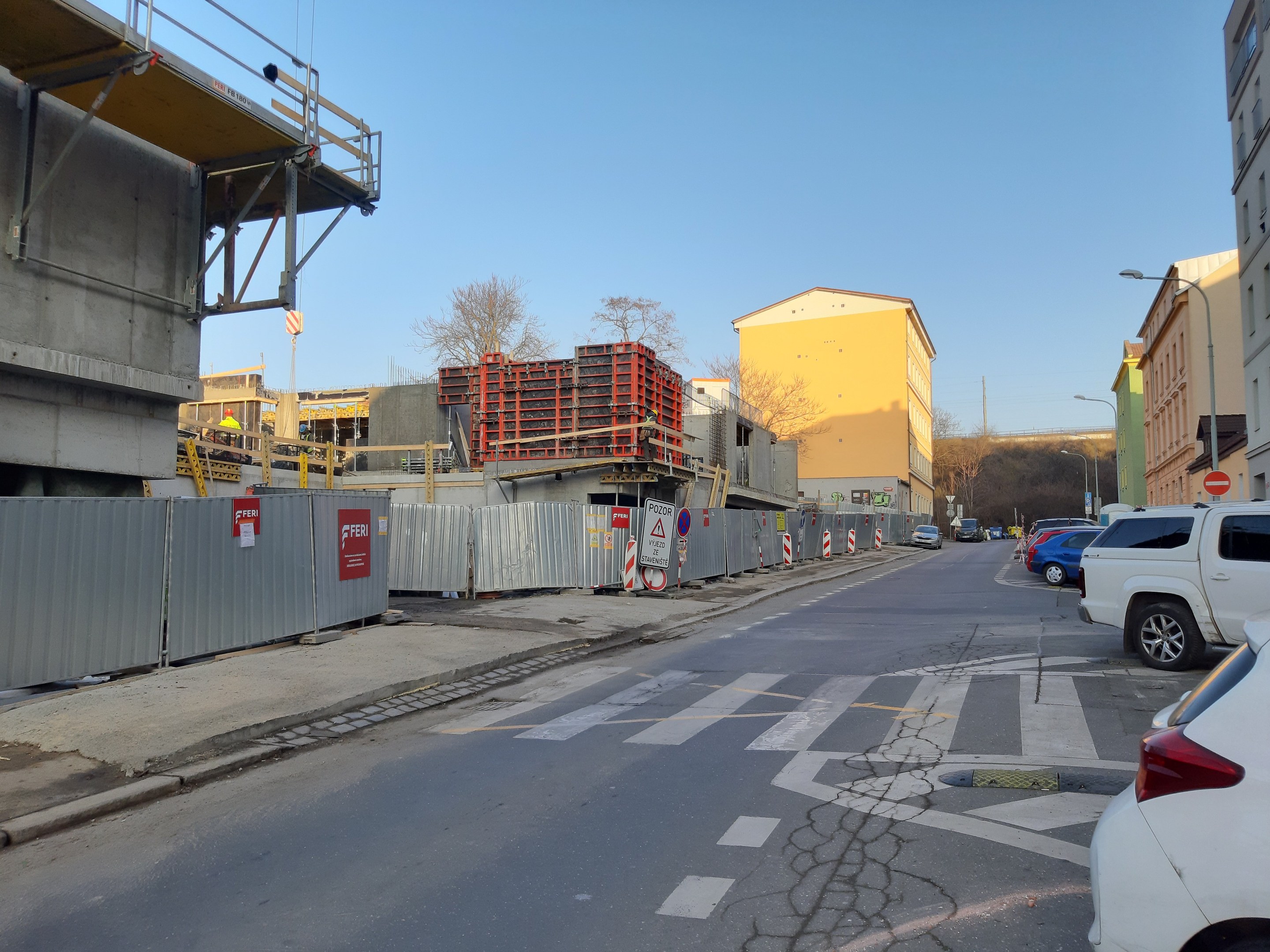 Stadtstraße mit parkenden Autos, Gebäuden, Laternen, Bäumen und Himmel im Hintergrund.