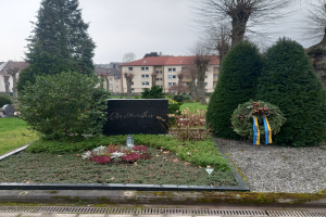Ein Denkmal für Opfer des Zweiten Weltkriegs steht in einem Park, geschmückt mit einem Kranz und umgeben von Grün, mit Gebäuden im Hintergrund.