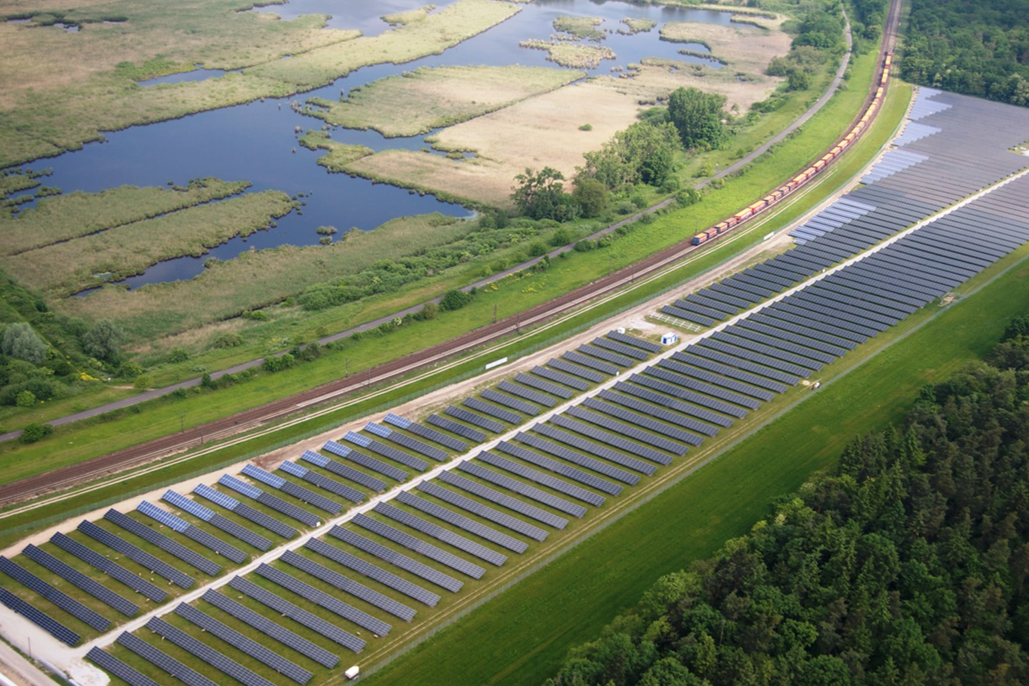 Luftaufnahme eines Solarparks mit Panelen in einem Feld, umgeben von Bäumen, Gras, Pflanzen und Wasser, mit einem Zug auf einem nahen Bahngeleis.