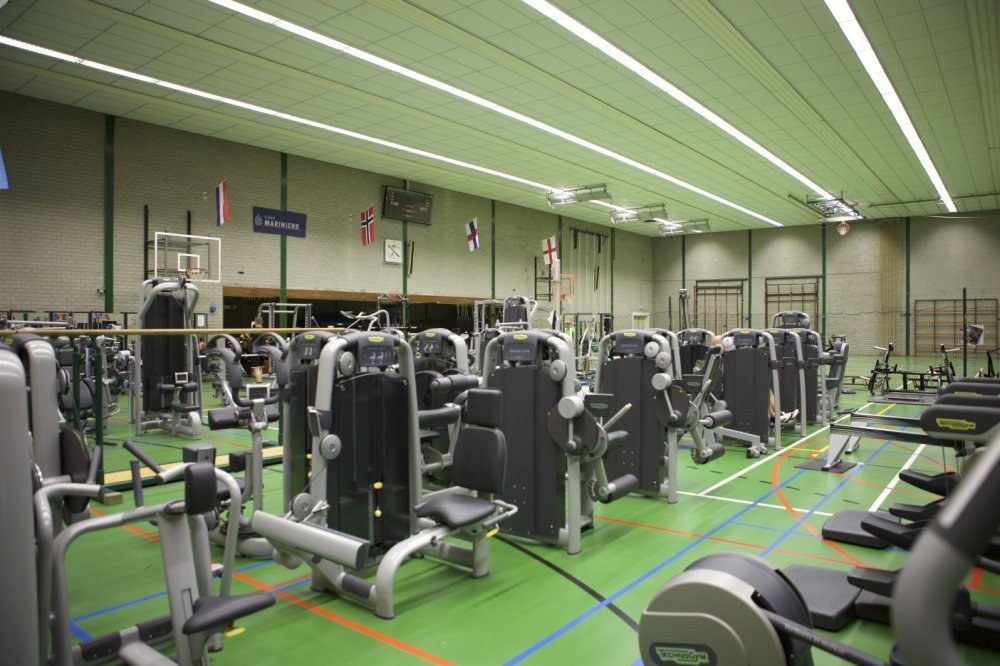 Ein ger%C3%A4umiges Fitnessstudio mit verschiedenen Trainingsger%C3%A4ten, einer Wand mit Flaggen und Deckenbeleuchtung.