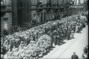 Schwarzes und weißes Foto einer großen Menge, die bei einer Parade marschiert, einige halten Gewehre und gehen an einem Gebäude mit einem Wasserzeichen in der oberen rechten Ecke vorbei.