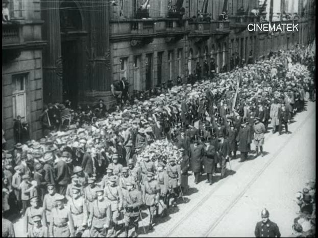 Schwarzes und weißes Foto einer großen Menge, die bei einer Parade marschiert, einige halten Gewehre und gehen an einem Gebäude mit einem Wasserzeichen in der oberen rechten Ecke vorbei.