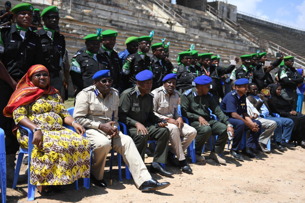 Eine Gruppe von Menschen, die auf Stühlen sitzen und einer Menge uniformierter Personen gegenüberstehen, einige halten Gegenstände, mit Treppen, Geländern und Pfählen im Hintergrund.