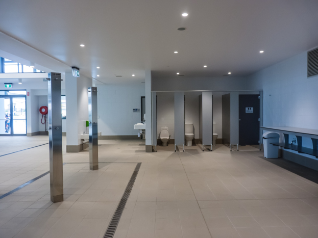 Eine große öffentliche Toilette mit mehreren Toiletten, Säulen, einem Mülleimer, einer Tür, einem Schild, einem Feuerlöscher und Deckenleuchten.