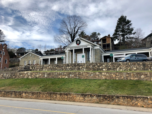 Steinwand mit geparkten Autos, angrenzend an ein Gebäude mit der Aufschrift "Welterbe-Museum", mit Fenstern, Türen, Bäumen und einem bewölkten Himmel.