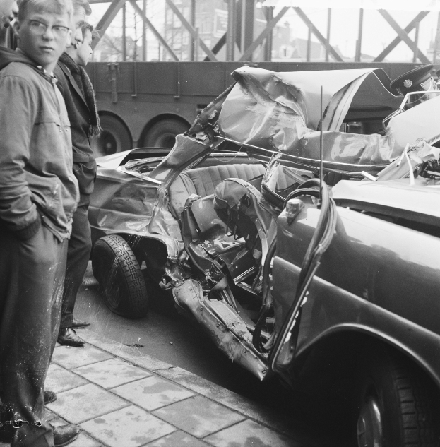 Schwarzes und weißes Bild eines verunglückten Autos am Straßenrand mit Menschen in der Nähe, einem Lastwagen und einer Brücke im Hintergrund.
