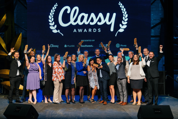 Gruppe von Menschen auf einer Bühne mit Auszeichnungen, mit Rednern darunter und einem Bildschirm im Hintergrund, der "Classy Awards 2019" anzeigt.