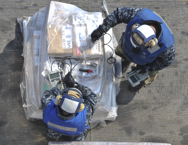 Zwei Männer in blauen Uniformen und Helmen arbeiten an einem großen Stück Ausrüstung, das wie ein Drohne aussieht, und halten verschiedene Gegenstände, mit einer Plastikabdeckung auf dem Boden.