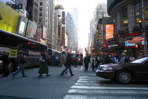 Eine belebte Stadtstraße mit Fahrzeugen, Fußgängern an einer Zebrastreifenfurt, Gebäuden, Schildern, Verkehrsampeln und einem blauen Himmel.