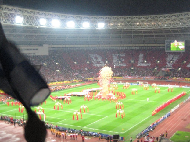 Ein Stadion mit Flutlicht, Eisengittern, Zuschauern, stehenden Personen, Werbetafeln, Anzeigetafeln, einer Überwachungskamera, Fahnen und Dekorationselementen.