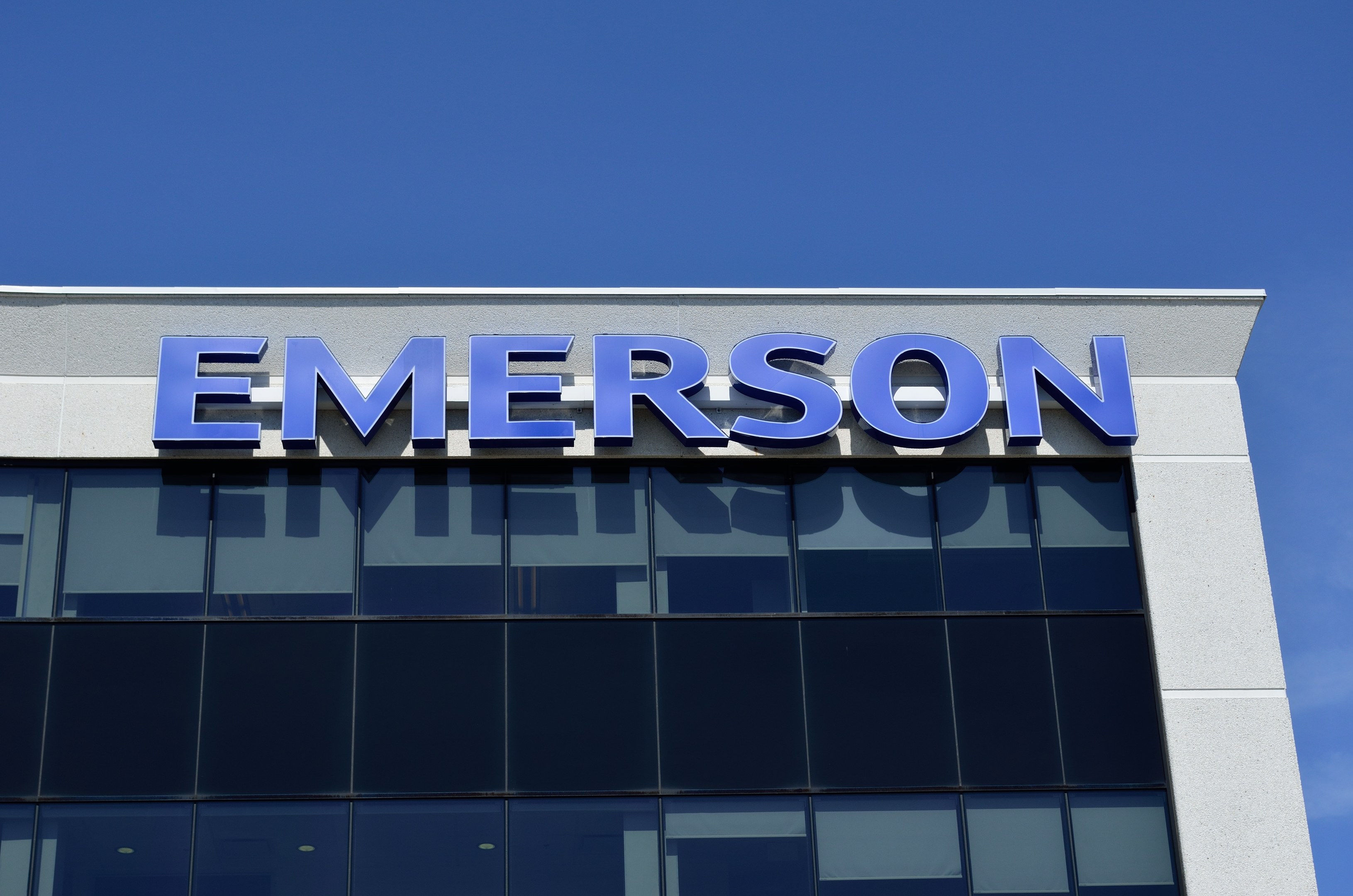 Emerson-Logo an einer Gebäudeseite mit dem Text "Emerson investiert 1,5 Milliarden USD in den USA" vor einem klaren blauen Himmel.