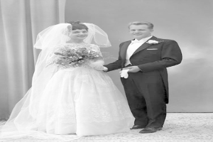 Ein Schwarz-Weiß-Foto eines lachenden Mannes und einer Frau, die an ihrem Hochzeitstag nebeneinanderstehen, wobei die Frau ein weißes Kleid trägt und einen Strauß hält und ein Vorhang und eine Wand im Hintergrund zu sehen sind.