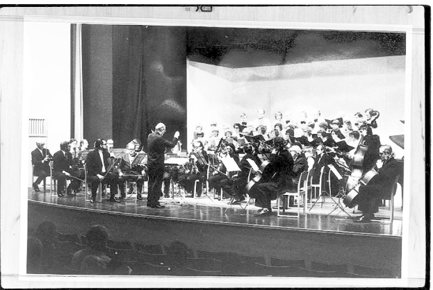 Ein Schwarz-Weiß-Foto eines Dirigenten in Anzug und Krawatte, der auf einer Bühne steht und ein Orchester mit Geigen, Celli und Flöten leitet, während das Publikum auf Stühlen aufmerksam zuhört.