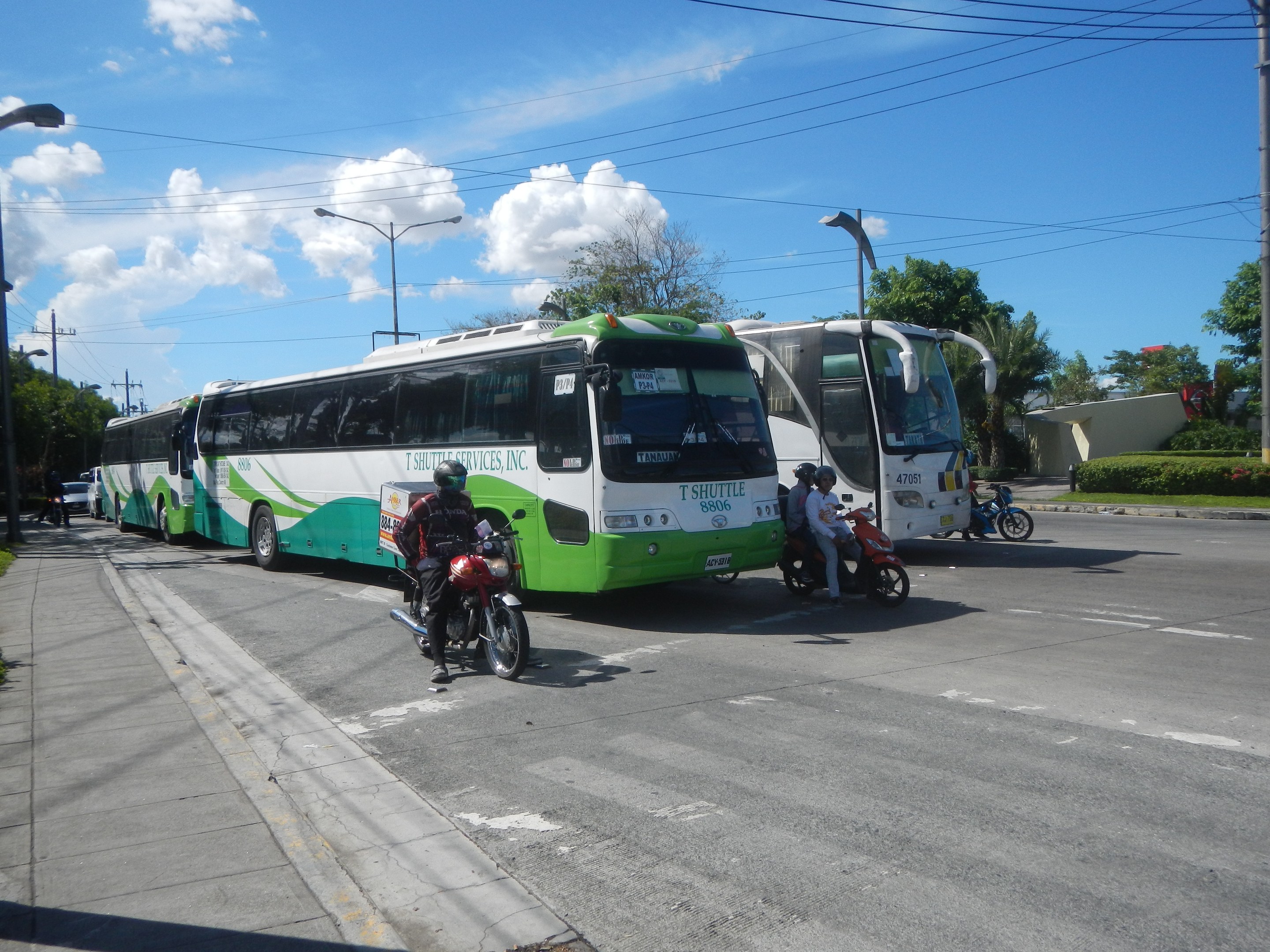 Ein grüner und weißer Shuttlebus parkt am Straßenrand mit Motorrädern davor, einem grasbewachsenen Fußweg links und Gebäuden, Bäumen und Laternen im Hintergrund unter einem klaren blauen Himmel.