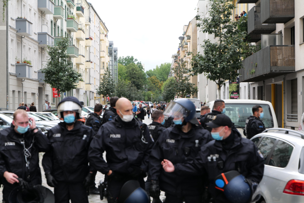 Polizisten in Masken und Helmen gehen eine Straße entlang, die von parkenden Autos gesäumt ist, flankiert von Gebäuden mit Balkonen und Geländern, mit Bäumen und einem klaren Himmel im Hintergrund.