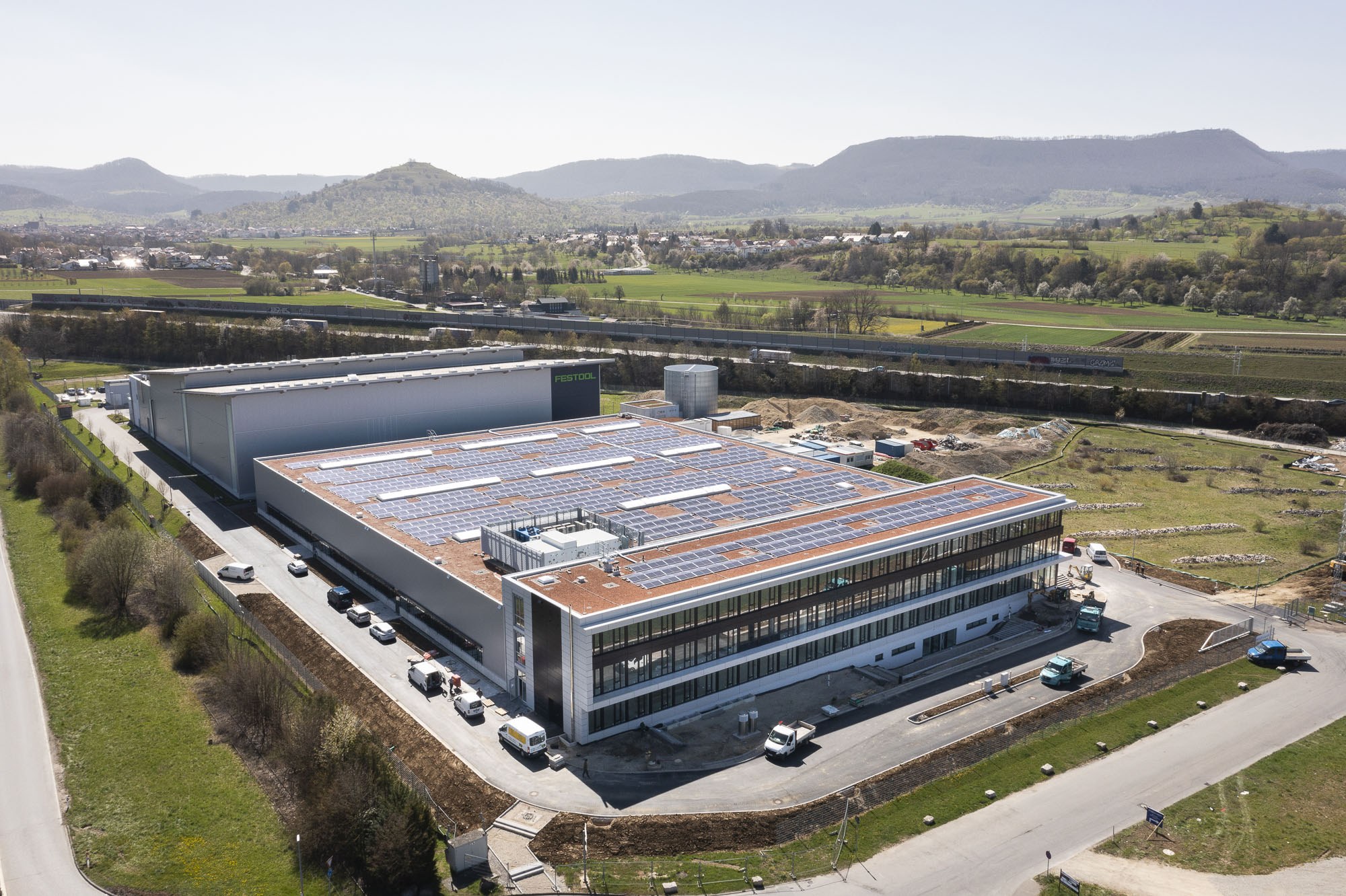 Luftaufnahme eines große industriellen Gebäudes mit Solarpanelen auf dem Dach, umgeben von Fahrzeugen, Bäumen, Gras und Strommasten, vor Hühgeln und einem klaren blauen Himmel.