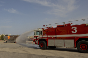 Rote Feuerwehrauto am Straßenrand geparkt, das Wasser auf den Boden sprüht mit Verkehrskegeln, Gebäuden, Bäumen und einem klaren blauen Himmel im Hintergrund.