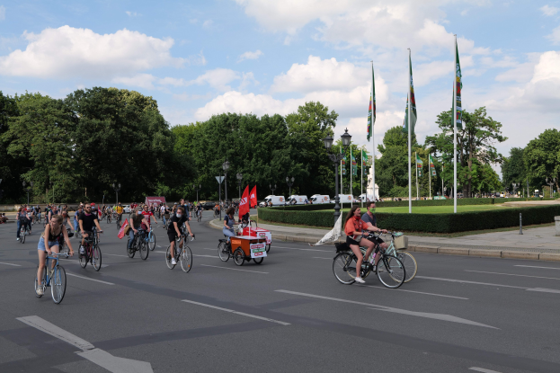 Eine Gruppe von Menschen, die auf Fahrrädern eine von Bäumen gesäumte Straße mit Laternenmasten, Fahnen und Fahrzeugen im Hintergrund entlangfahren, während der Pariser Radtour 2016.