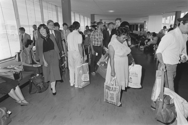 Ein Schwarz-Weiß-Bild von Reisenden in einem Flughafen, einige sitzen auf Bänken und andere stehen, mit einem Mann und einer Frau, die Taschen halten, in der Mitte und Fensterjalousien auf der linken Seite.