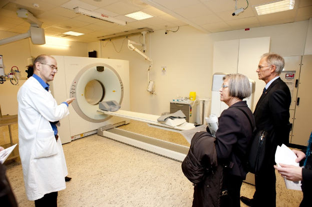 Arzt in weißem Kittel erklärt eine CT-Scan-Maschine zwei Menschen in schwarzen Mänteln.