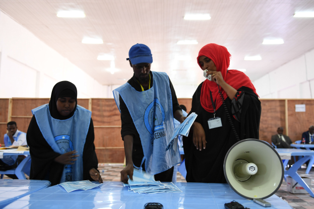 Eine Gruppe von Menschen versammelt sich um einen Tisch mit einem Megafon, Papieren und anderen Gegenständen an einer Wahllokal, während andere im Hintergrund auf Stühlen sitzen und stehen, unter Deckenlampen.