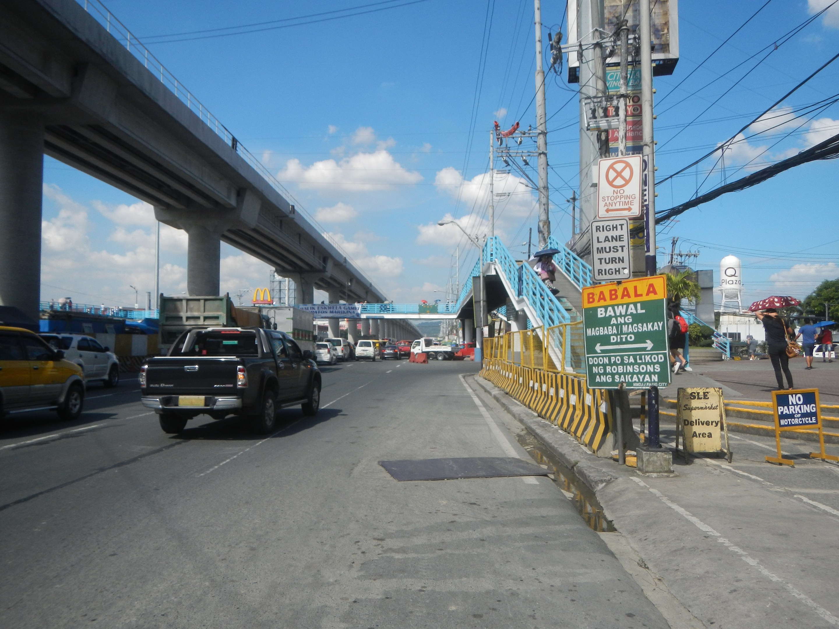 Eine belebte Straße mit Fahrzeugen, eine Brücke, Strommasten, Schilder, Fußgänger, Bäume, Gebäude und einen bewölkten Himmel.