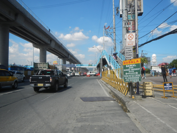 Eine belebte Straße mit Fahrzeugen, eine Brücke, Strommasten, Schilder, Fußgänger, Bäume, Gebäude und einen bewölkten Himmel.
