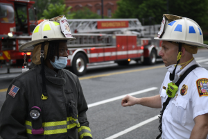 Zwei Feuerwehrleute in Helmen und Gesichtsmasken unterhalten sich auf einer Straße mit Fahrzeugen, Bäumen, Gebäuden und einem Pfahl im Hintergrund.