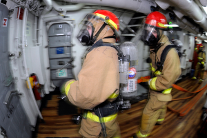 Zwei Feuerwehrleute in Schutzausrüstung, darunter Helme und Gasmasken, gehen durch das Innere eines Flugzeugs, mit einer Tür auf der linken Seite und Rohren und anderen Gegenständen auf der rechten Seite.