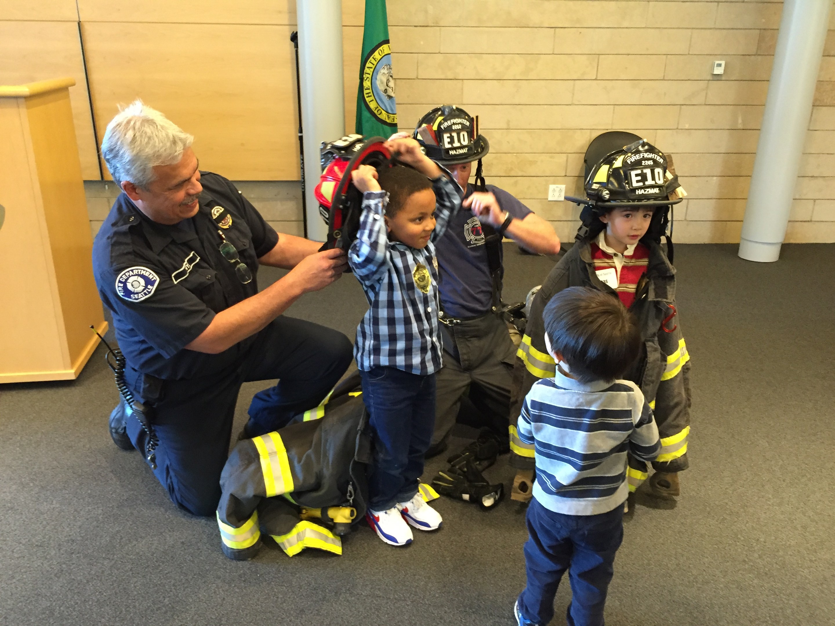 Eine Gruppe von Kindern in Feuerwehruniformen und ein Feuerwehrmann in Uniform stehen um den Feuerwehrmann herum, der einen Helm hält, mit einer Tasche auf dem Boden in einer Feuerwache.