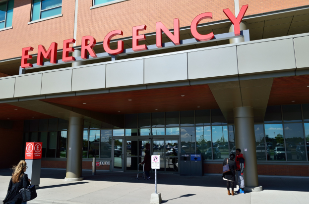 Eine Gruppe von Menschen geht vor einem Notfallgebäude mit Glaswänden und Säulen, einem Namensschild, Hinweistafeln auf Pfählen und einem Abfalleimer.