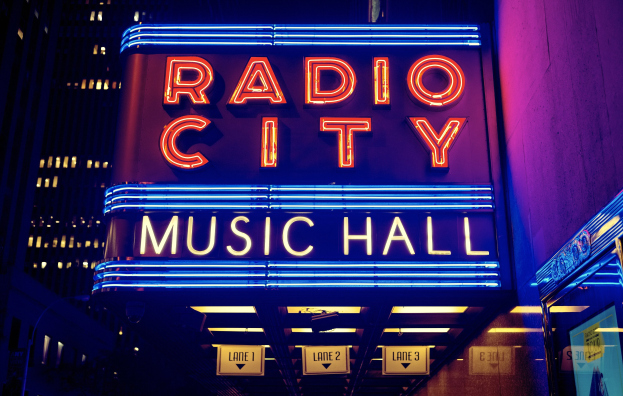 Das erleuchtete Radio City Music Hall in New York City mit hohen Gebäuden im Hintergrund, das eine 'Radio City'-Leuchtschrift und mehrere kleinere Schilder auf der rechten Seite zeigt.