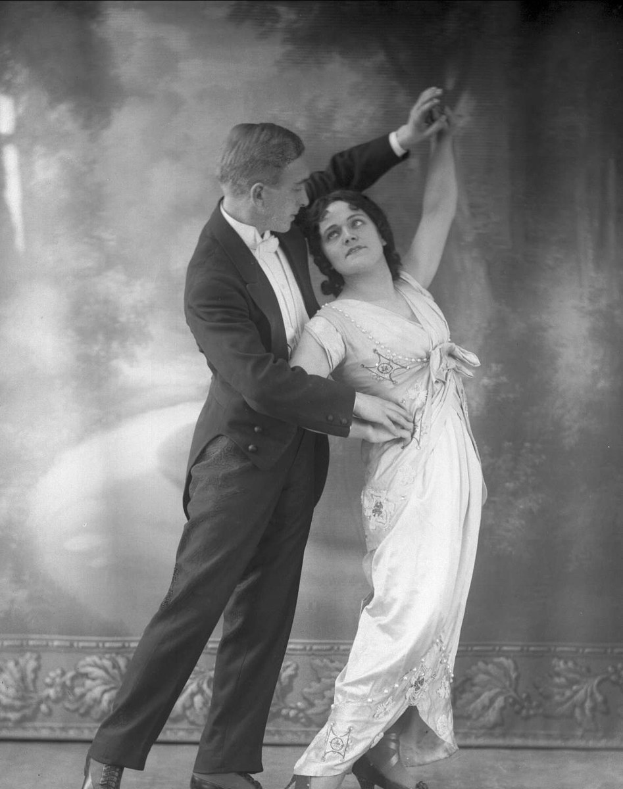 Schwarzes und weißes Foto eines Mannes in einem Anzug und einer Frau, die Tango tanzen, mit kunstvollen Wanddesigns im Hintergrund.