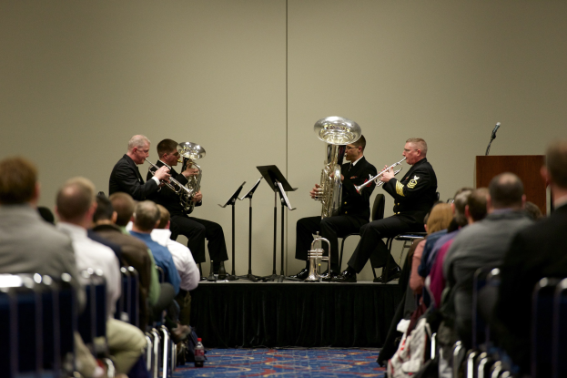 Gruppe von Männern, die auf einer Bühne Brass-Instrumente spielen, mit Notenständern, einem Podium und einem Mikrofon, vor einem sitzenden Publikum und einer Wand im Hintergrund.