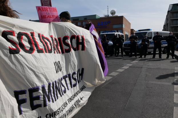 Eine Gruppe von Menschen marschiert auf einer Straße, hält eine Fahne mit der Aufschrift 'Solidarität und Feminismus', mit parkenden Fahrzeugen und Gebäuden im Hintergrund unter einem klaren blauen Himmel.