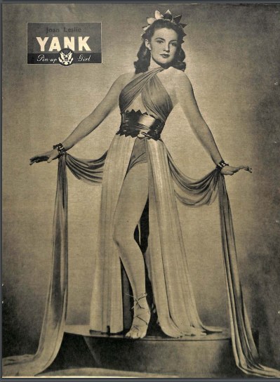 Ein Schwarz-Weiß-Foto der Schauspielerin Joan Leslie, die auf einer Bühne steht und ein langärmliges, hochgeschlossenes Kleid trägt, mit einem klassischen 1940er-Haarschnitt und dezentem Make-up, eingerahmt mit ihrem Namen oben drauf.